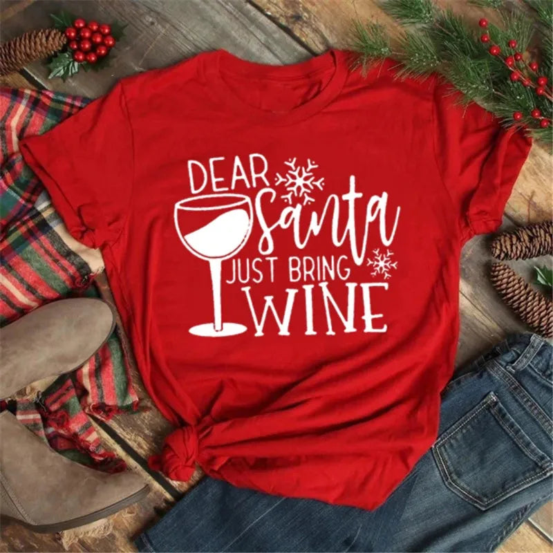 Maglietta rossa natalizia con scritta "Dear Santa Just Bring Wine", decorata con fiocchi di neve, su sfondo di legno con rami di pino e bacche rosse.
