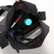 Borsa a tracolla nera aperta, contenente bottiglia d'acqua, ombrello, libro e penna. Design compatto, ideale per viaggi e uso quotidiano.