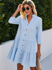 Donna con abito estivo a quadri azzurri, maniche a tre quarti, bottoni frontali, occhiali da sole, sfondo verde. Moda casual femminile.