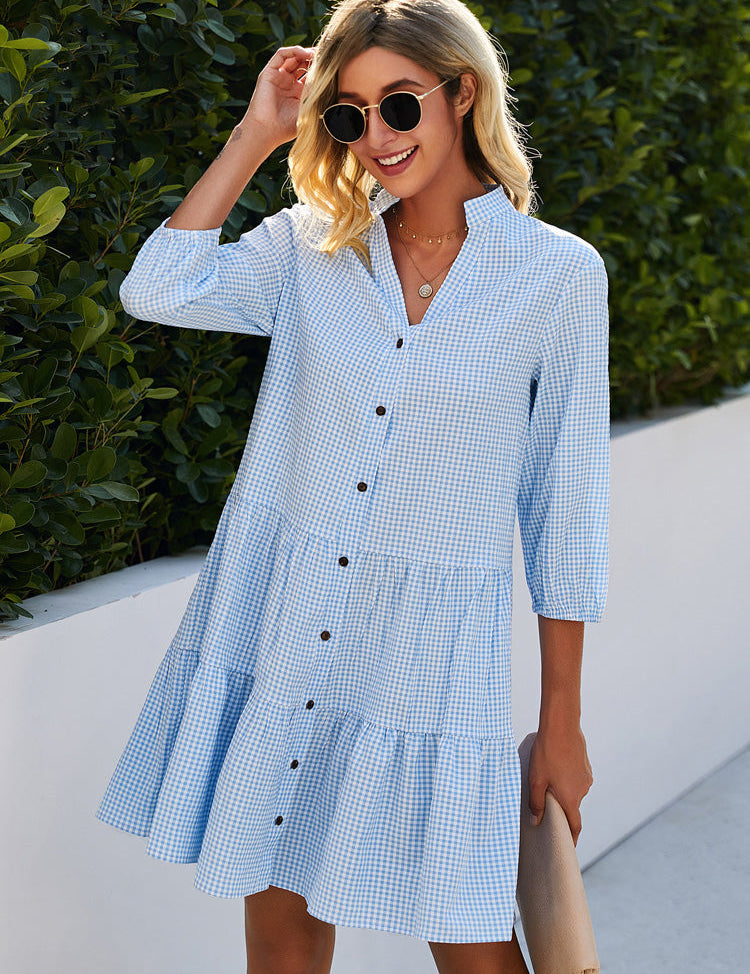 Donna con abito estivo a quadri azzurri, maniche a tre quarti, bottoni frontali, occhiali da sole, sfondo verde. Moda casual femminile.