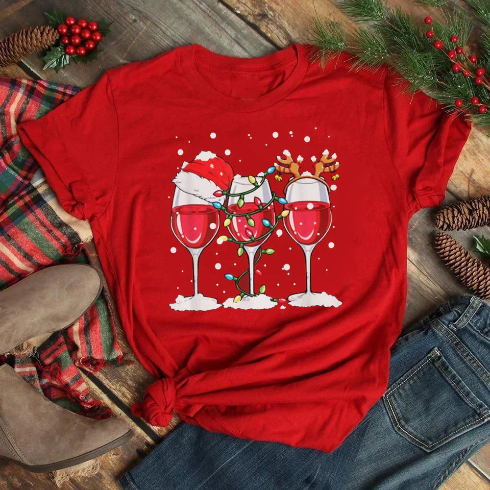 Maglietta rossa natalizia con tre bicchieri di vino decorati con cappello di Babbo Natale, luci e corna di renna, su sfondo di pigne e rami di pino.