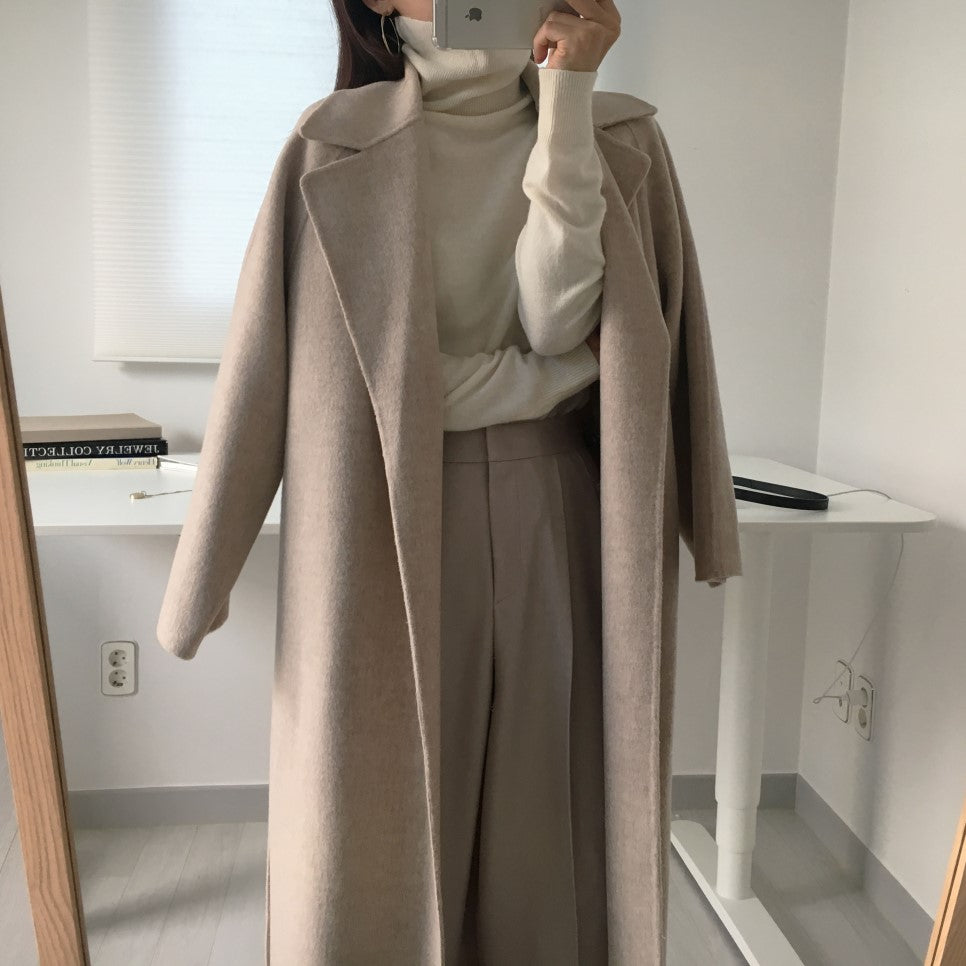 Cappotto lungo beige elegante, abbinato a dolcevita bianco e pantaloni larghi, in una stanza moderna. Moda autunnale, abbigliamento femminile chic.