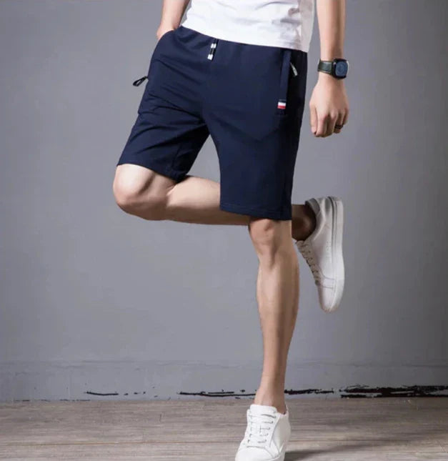 Pantaloncini sportivi blu da uomo, tasche con zip, indossati con t-shirt bianca e scarpe da ginnastica bianche. Stile casual estivo.