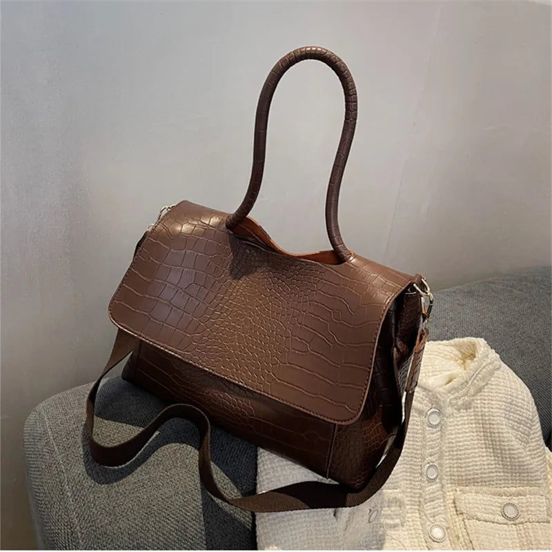 Borsa a tracolla in pelle marrone con texture coccodrillo, manico lungo, su divano grigio. Elegante accessorio moda donna.