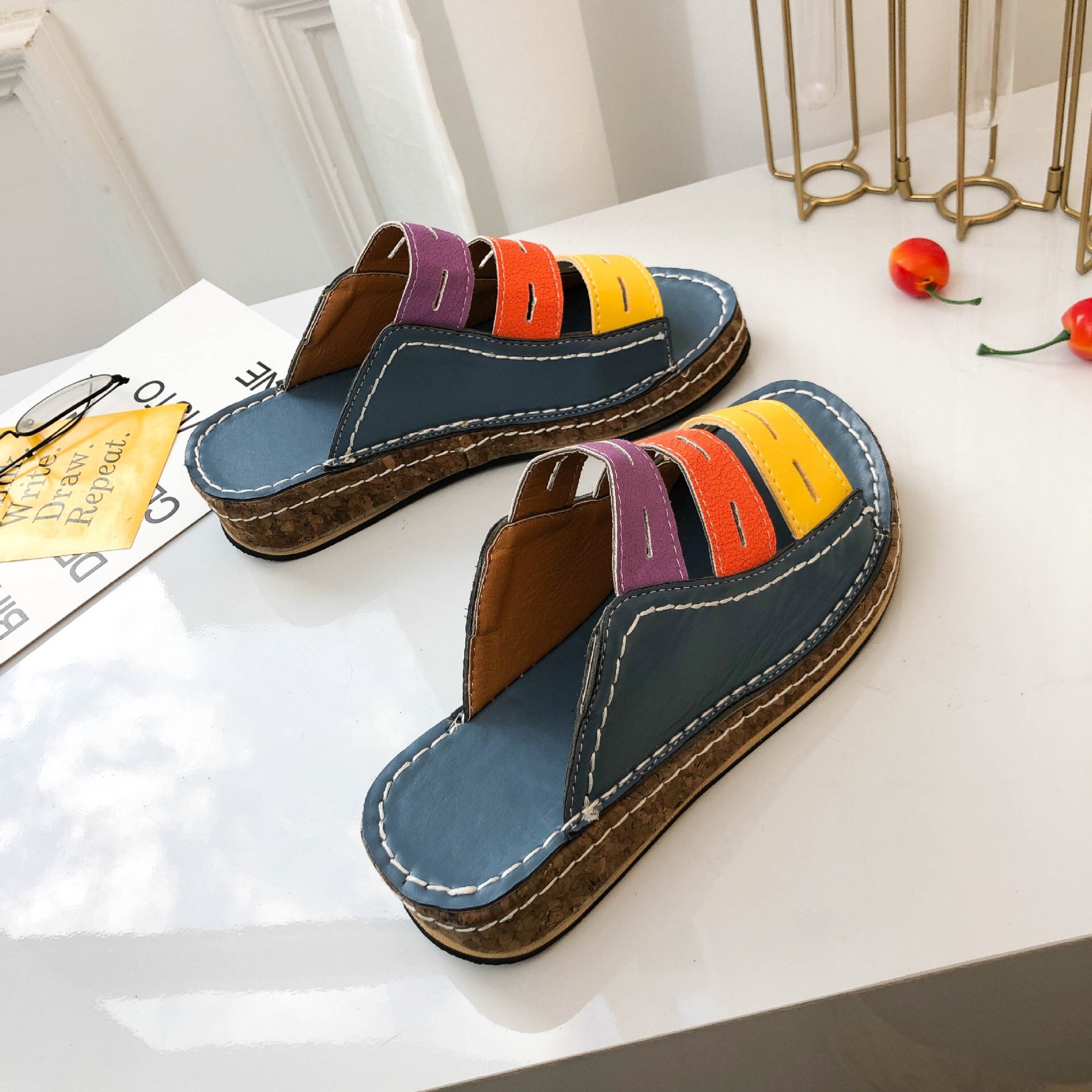 Sandali donna multicolore con suola in sughero, design a fasce blu, viola, arancione e giallo, su tavolo bianco. Scarpe estive moda casual.