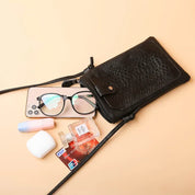 Borsa a tracolla in pelle nera con occhiali, smartphone, carta di credito, auricolari e disinfettante su sfondo beige. Accessori moda eleganti.