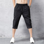 Pantaloni corti sportivi neri da uomo, lunghezza al polpaccio, con elastico in vita e tasche laterali, indossati con scarpe da ginnastica bianche.