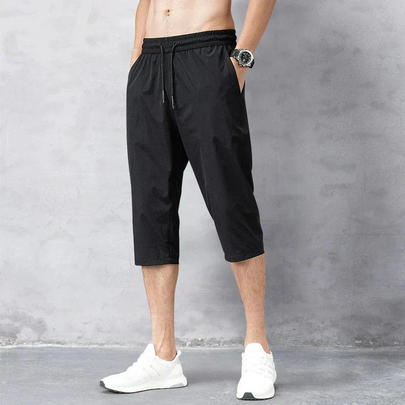 Pantaloni sportivi neri da uomo, lunghezza tre quarti, con elastico in vita e tasche laterali. Indossati con scarpe da ginnastica bianche.