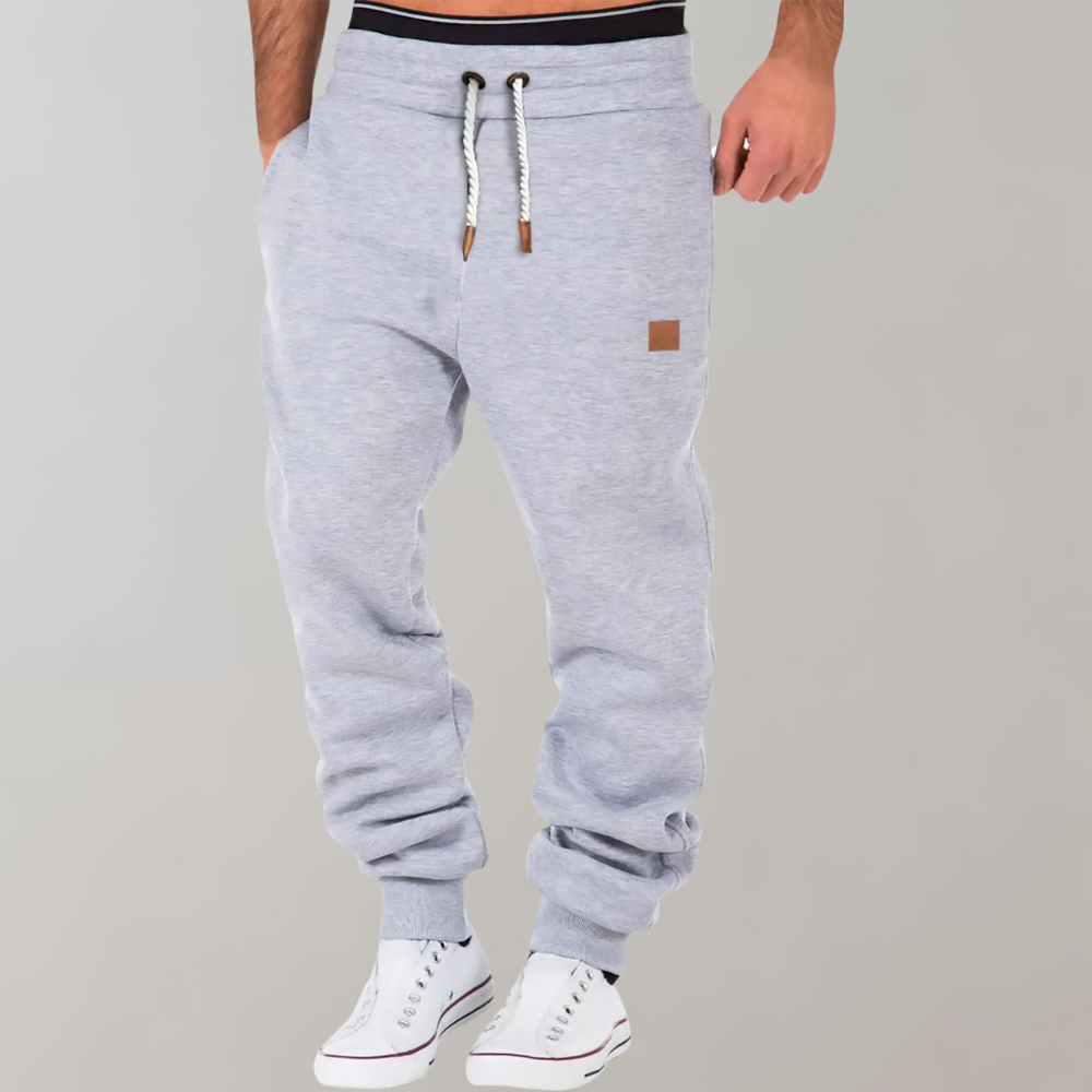 Pantaloni tuta grigi da uomo con coulisse, tasche laterali, polsini elasticizzati, abbinati a scarpe da ginnastica bianche.
