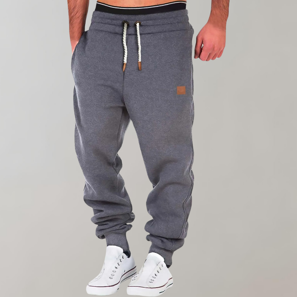 Pantaloni tuta grigi da uomo con coulisse, tasche laterali e polsini elasticizzati, abbinati a scarpe da ginnastica bianche.