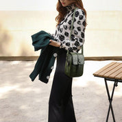 Donna con camicia a fiori bianca e nera, pantaloni neri, borsa a tracolla verde, giacca verde in mano, accanto a tavolo in legno. Moda autunnale elegante.
