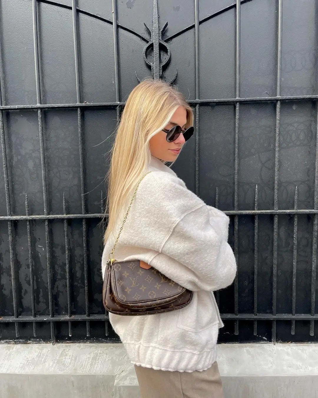 Donna con occhiali da sole indossa giacca beige e borsa a tracolla marrone con logo, davanti a cancello nero. Moda autunnale, stile urbano.
