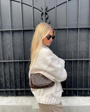 Donna con occhiali da sole indossa giacca beige e borsa a tracolla marrone con logo, davanti a cancello nero. Moda autunnale, stile urbano.