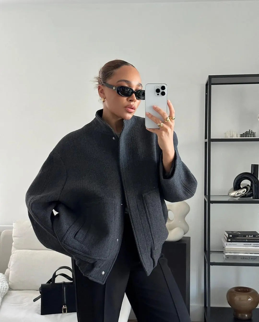 Donna con occhiali da sole indossa giacca grigia oversize, pantaloni neri, scatta selfie con iPhone in salotto moderno. Moda autunnale elegante.