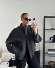 Donna con occhiali da sole indossa giacca grigia oversize, pantaloni neri, scatta selfie con iPhone in salotto moderno. Moda autunnale elegante.
