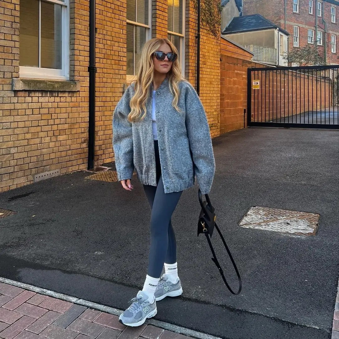 Donna con occhiali da sole indossa giacca grigia oversize, leggings blu scuro e scarpe da ginnastica grigie, cammina su strada asfaltata.