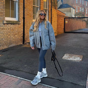 Donna con occhiali da sole indossa giacca grigia oversize, leggings blu scuro e scarpe da ginnastica grigie, cammina su strada asfaltata.