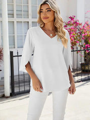 Donna con capelli biondi indossa blusa bianca a maniche lunghe e pantaloni abbinati, davanti a una casa con fiori rosa. Moda elegante e casual.