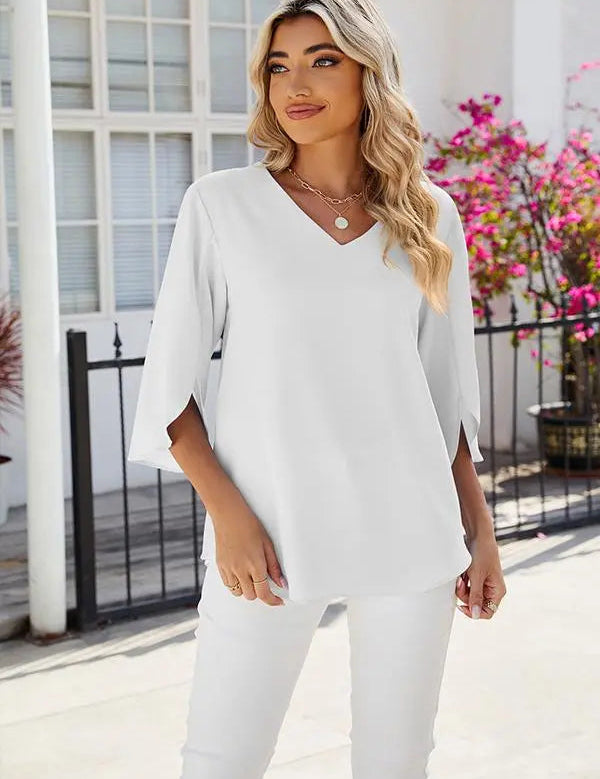 Donna con capelli biondi indossa blusa bianca a maniche lunghe e pantaloni abbinati, davanti a una casa con fiori rosa. Moda elegante e casual.