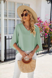 Donna con cappello di paglia, occhiali da sole e borsa intrecciata, indossa blusa verde chiaro e pantaloni bianchi. Sfondo con fiori e finestra.