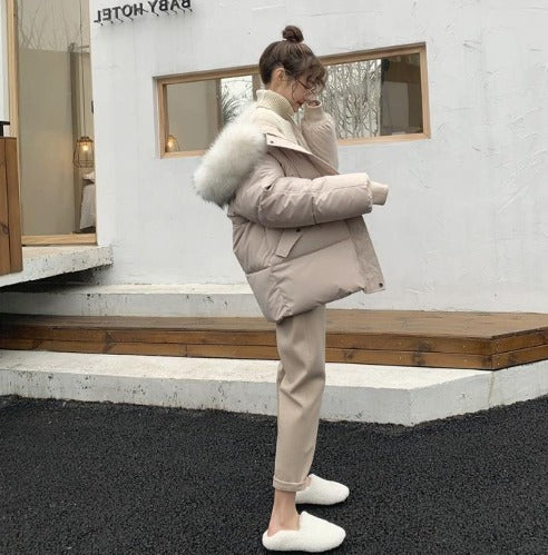 Donna con cappotto invernale beige con cappuccio in pelliccia, pantaloni coordinati e pantofole bianche, all'esterno di un edificio moderno.