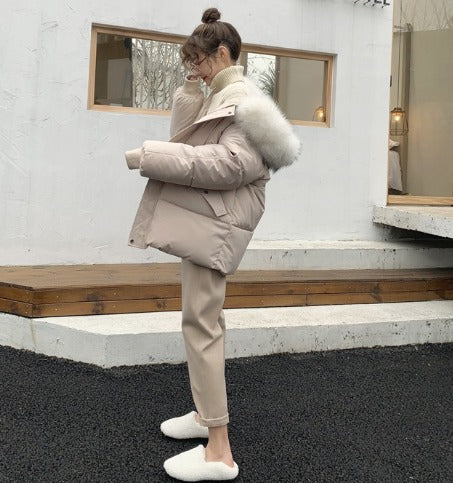 Donna con giacca imbottita beige con cappuccio in pelliccia, pantaloni chiari e pantofole bianche, all'aperto su sfondo di edificio moderno.