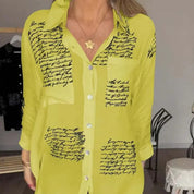 Camicia donna gialla con scritte nere, maniche lunghe, colletto classico, bottoni frontali, tasca sul petto, stile casual elegante.