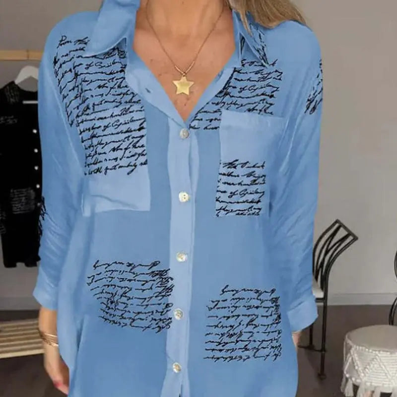 Camicia donna azzurra con stampa scritte nere, colletto classico, bottoni frontali, tasca sul petto, maniche lunghe. Moda casual elegante.