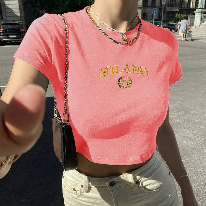 Donna con t-shirt rosa con scritta "Milano", catena dorata e borsa a tracolla, in strada soleggiata. Moda estiva, abbigliamento casual.