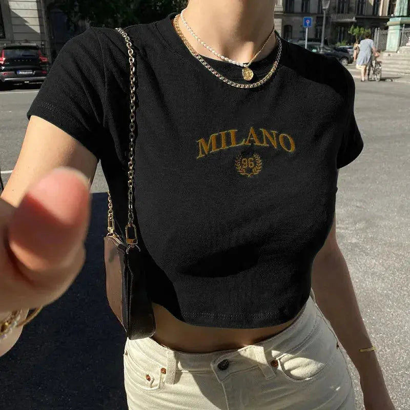 Donna con t-shirt nera "Milano", catena dorata, borsa a tracolla marrone, jeans chiari. Moda urbana estiva, stile casual chic.