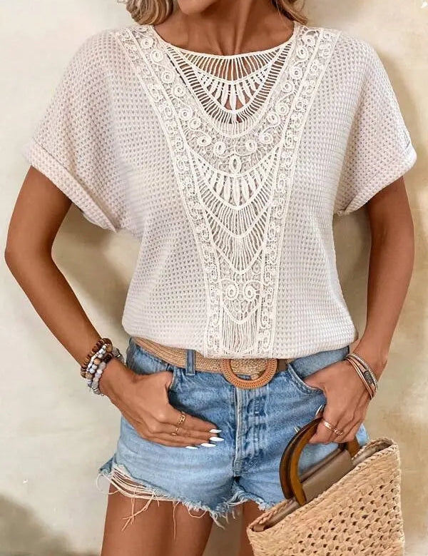 Donna con maglia bianca traforata e dettagli in pizzo, shorts di jeans, cintura marrone e borsa di paglia. Look estivo casual chic.