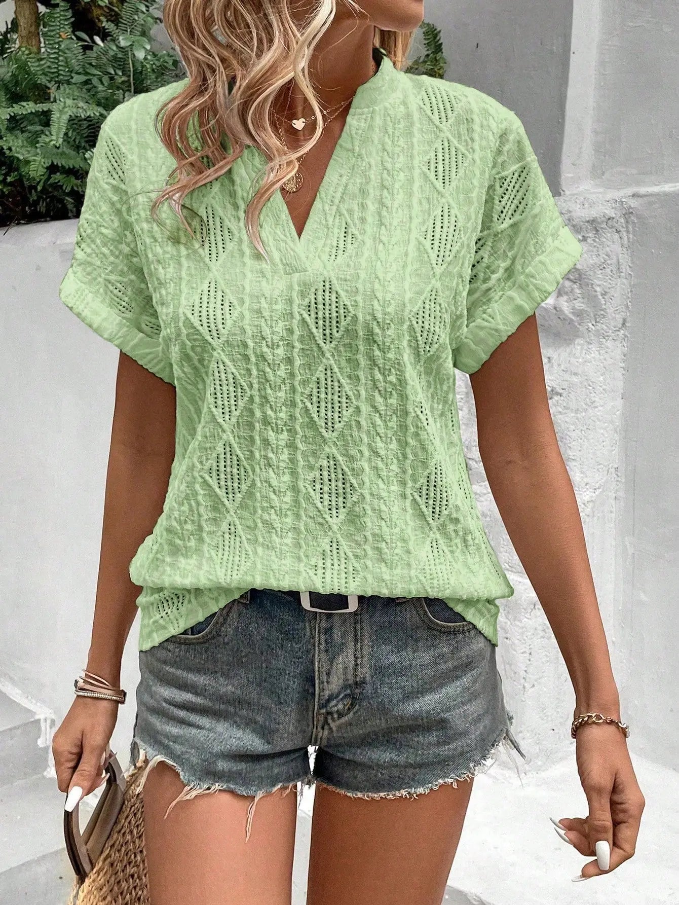 Donna con camicia verde ricamata e pantaloncini di jeans, stile estivo casual. Accessori: collana e borsa di paglia. Moda femminile trendy.