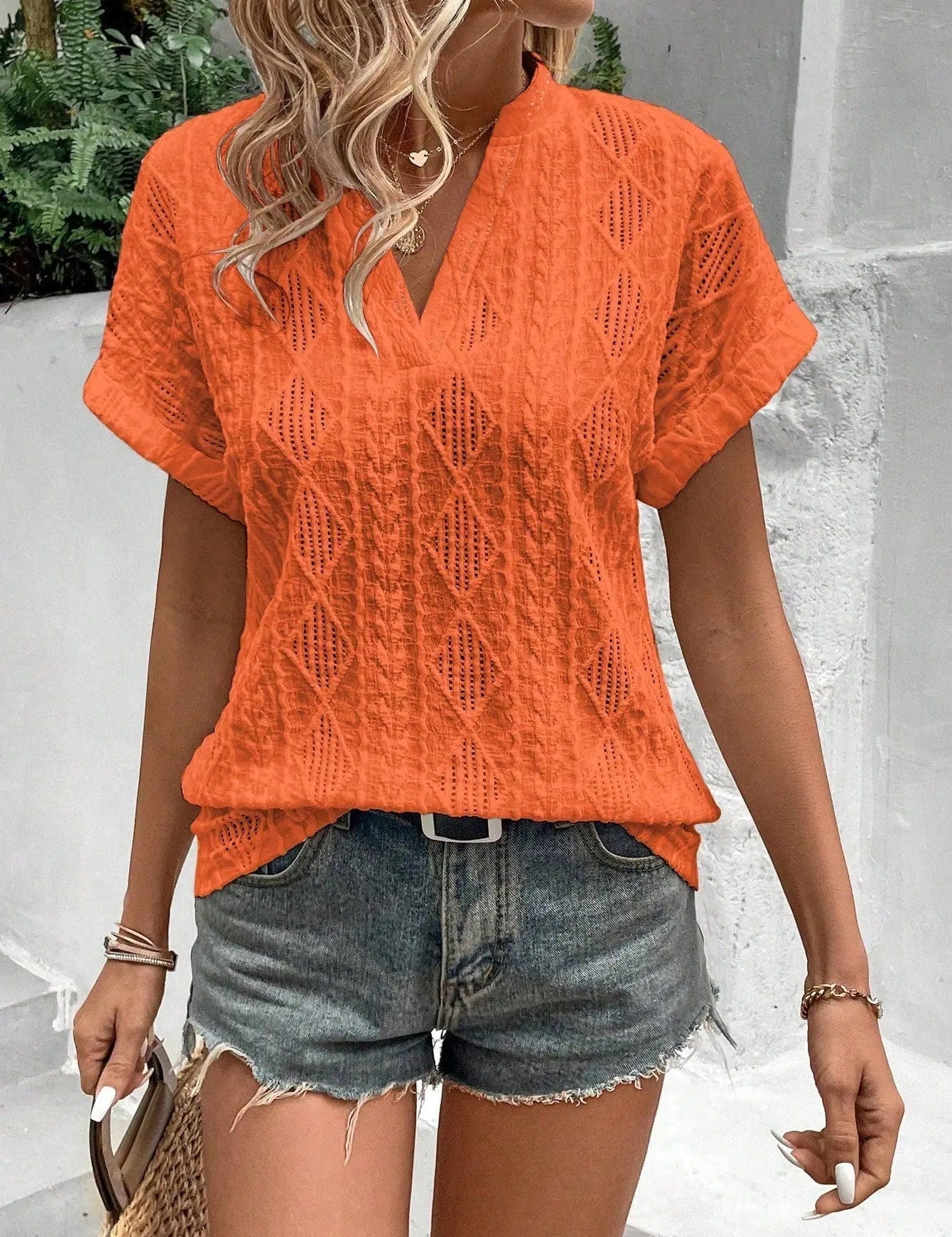 Donna con maglietta arancione traforata e pantaloncini di jeans, accessoriata con borsa di paglia. Moda estiva casual, look trendy e fresco.