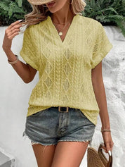 Donna con blusa gialla traforata, scollo a V, abbinata a shorts di jeans. Look estivo casual, accessori oro e cappello di paglia.