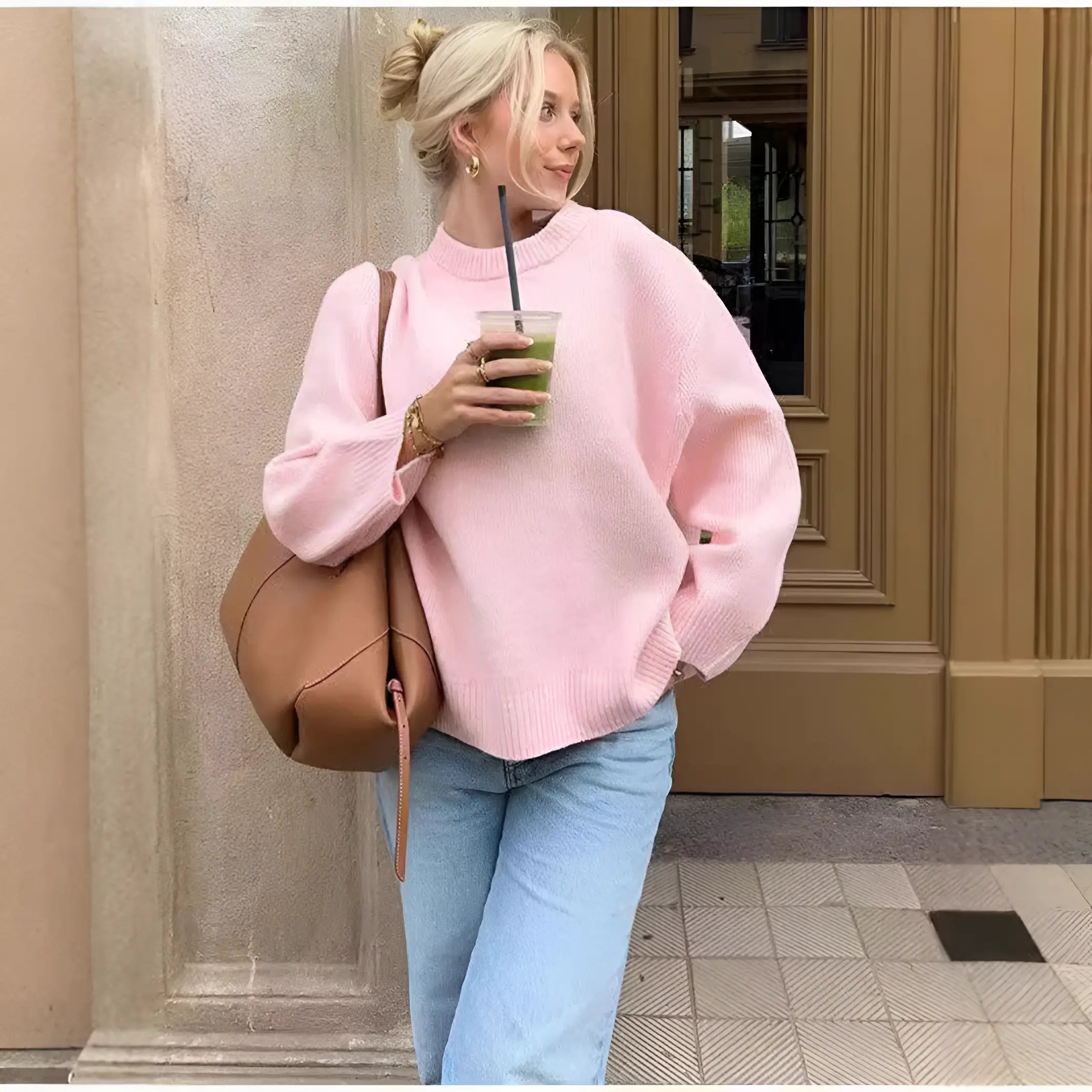 Bionda con maglione rosa e jeans chiari, sorseggia una bevanda verde. Borsa a tracolla marrone, sfondo urbano. Moda casual autunnale.