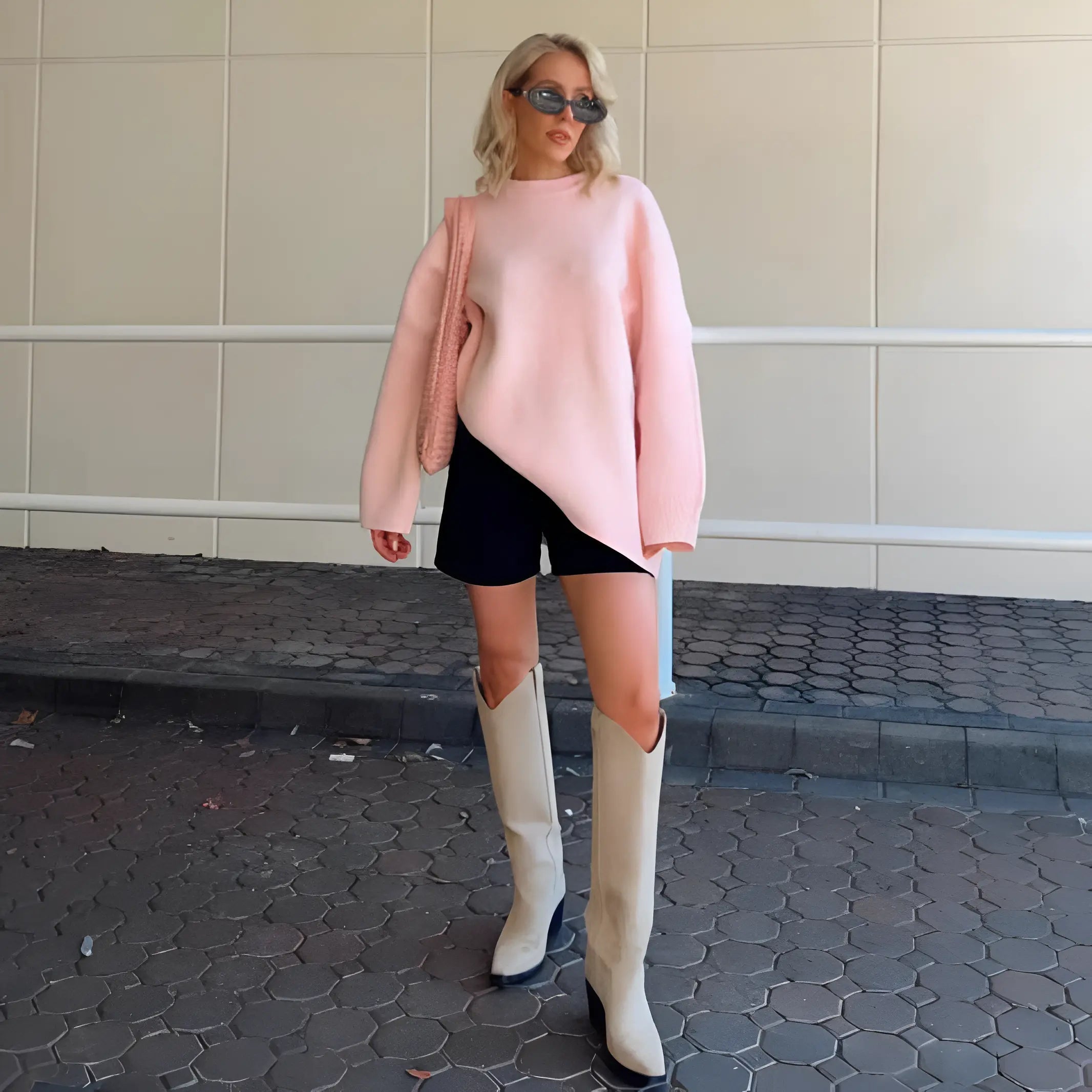 Donna con maglione rosa asimmetrico, shorts neri e stivali alti beige. Moda autunnale, occhiali da sole, borsa a tracolla. Look casual chic.