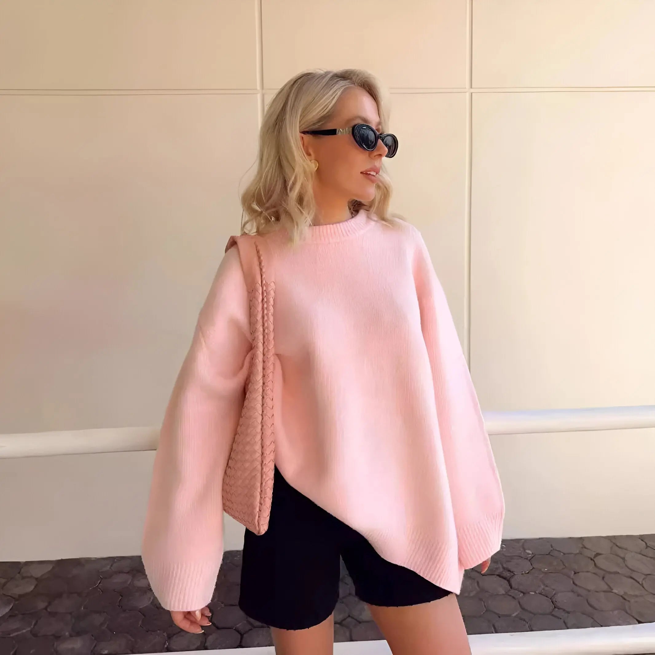 Donna con occhiali da sole indossa maglione rosa oversize e pantaloncini neri, borsa a tracolla rosa. Moda casual autunnale, stile urbano chic.