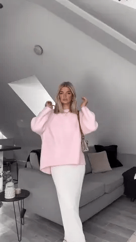 Donna con maglione rosa oversize e gonna bianca in soggiorno moderno. Moda autunnale, abbigliamento casual chic, arredamento minimalista.