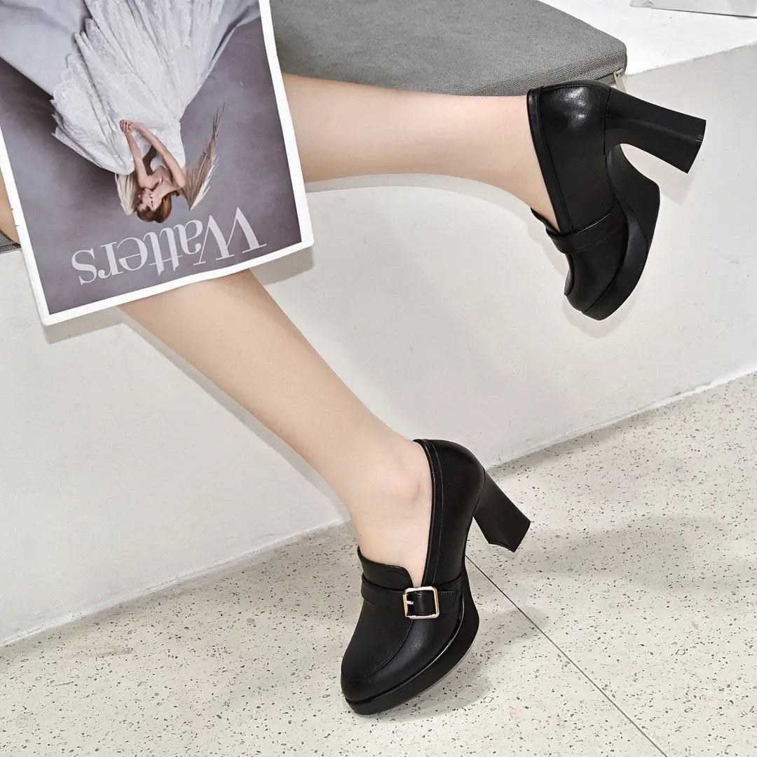 Scarpe décolleté nere con tacco alto e fibbia dorata, indossate da una persona seduta, con una rivista in mano. Eleganza e stile moderno.