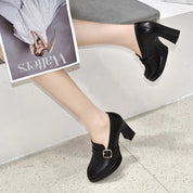 Scarpe décolleté nere con tacco alto e fibbia dorata, indossate da una persona seduta, con una rivista in mano. Eleganza e stile moderno.