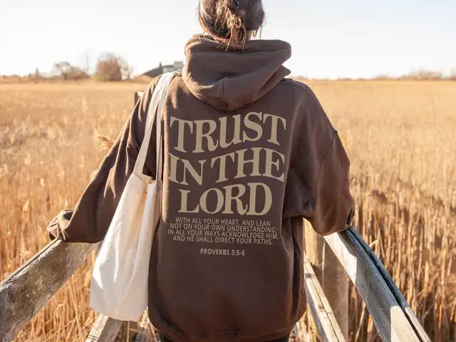 Donna con felpa marrone "Trust in the Lord", borsa a tracolla bianca, su passerella in legno in un campo di grano. Moda casual autunnale.