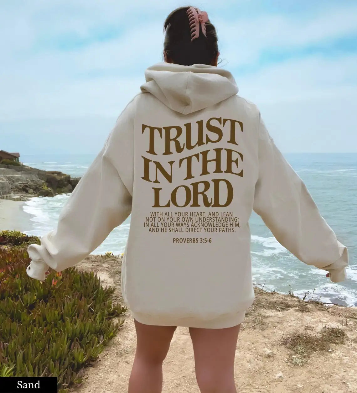 Donna in felpa beige con scritta "Trust in the Lord", vista mare. Abbigliamento casual, moda ispirata alla fede, paesaggio costiero.