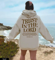 Donna in felpa beige con scritta "Trust in the Lord", vista mare. Abbigliamento casual, moda ispirata alla fede, paesaggio costiero.