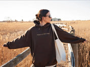 Donna con felpa marrone "Trust in the Lord", occhiali da sole, borsa bianca, su passerella in campagna. Moda casual autunnale, stile rilassato.