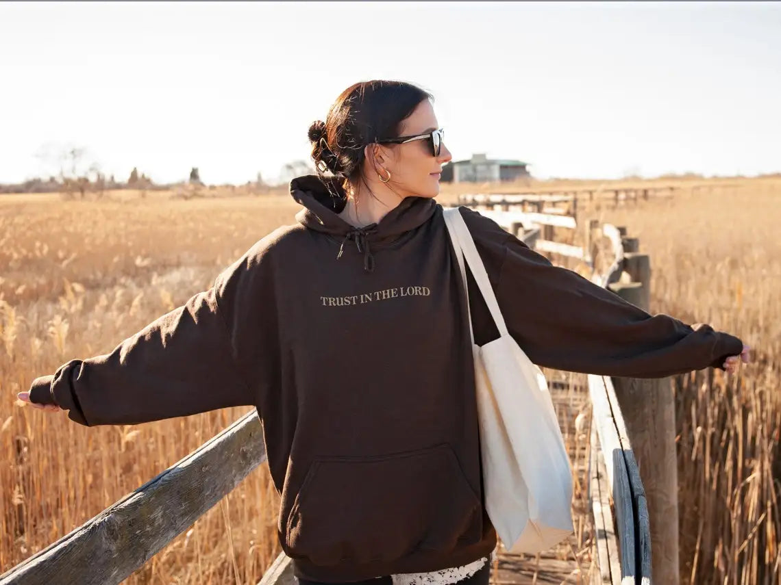 Donna con felpa marrone "Trust in the Lord", occhiali da sole, borsa bianca, su passerella in campagna. Moda casual autunnale, stile rilassato.