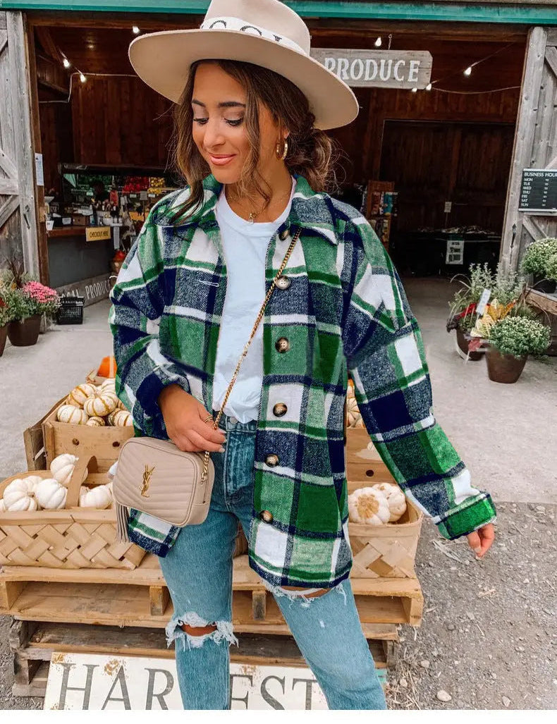 Donna con cappello beige e camicia a quadri verde e blu, davanti a un mercato agricolo con zucche. Moda autunnale, stile casual chic.
