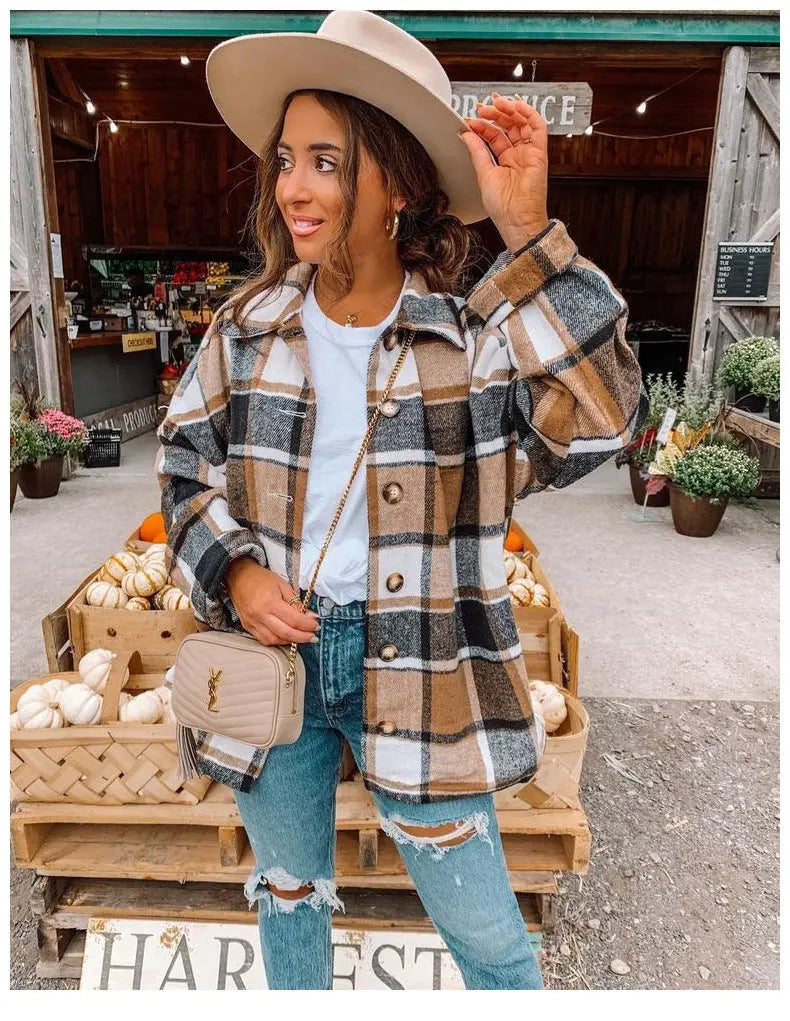 Donna con cappello beige e giacca a quadri marrone, bianca e nera, davanti a un negozio di campagna con zucche e fiori. Moda autunnale casual.