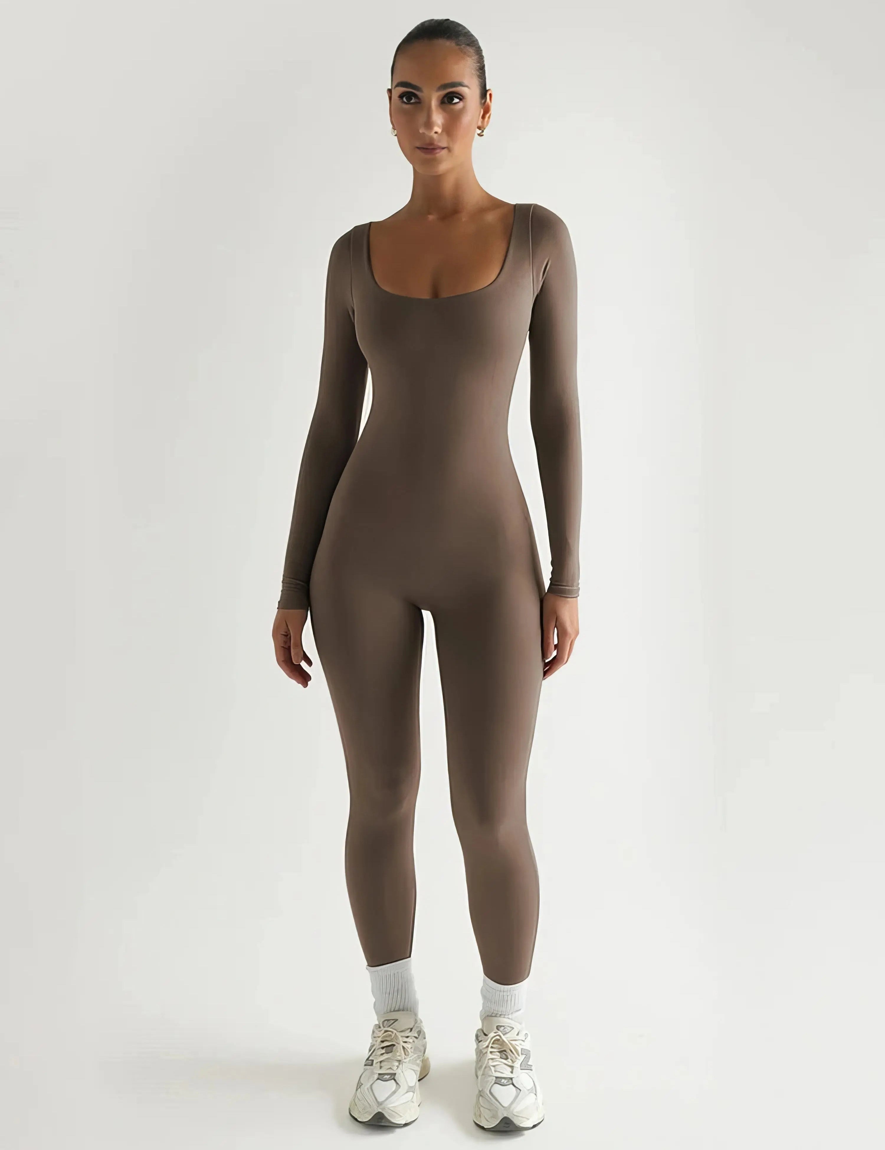 Donna in tuta aderente marrone, maniche lunghe, scarpe da ginnastica bianche, sfondo bianco. Abbigliamento sportivo elegante e moderno.