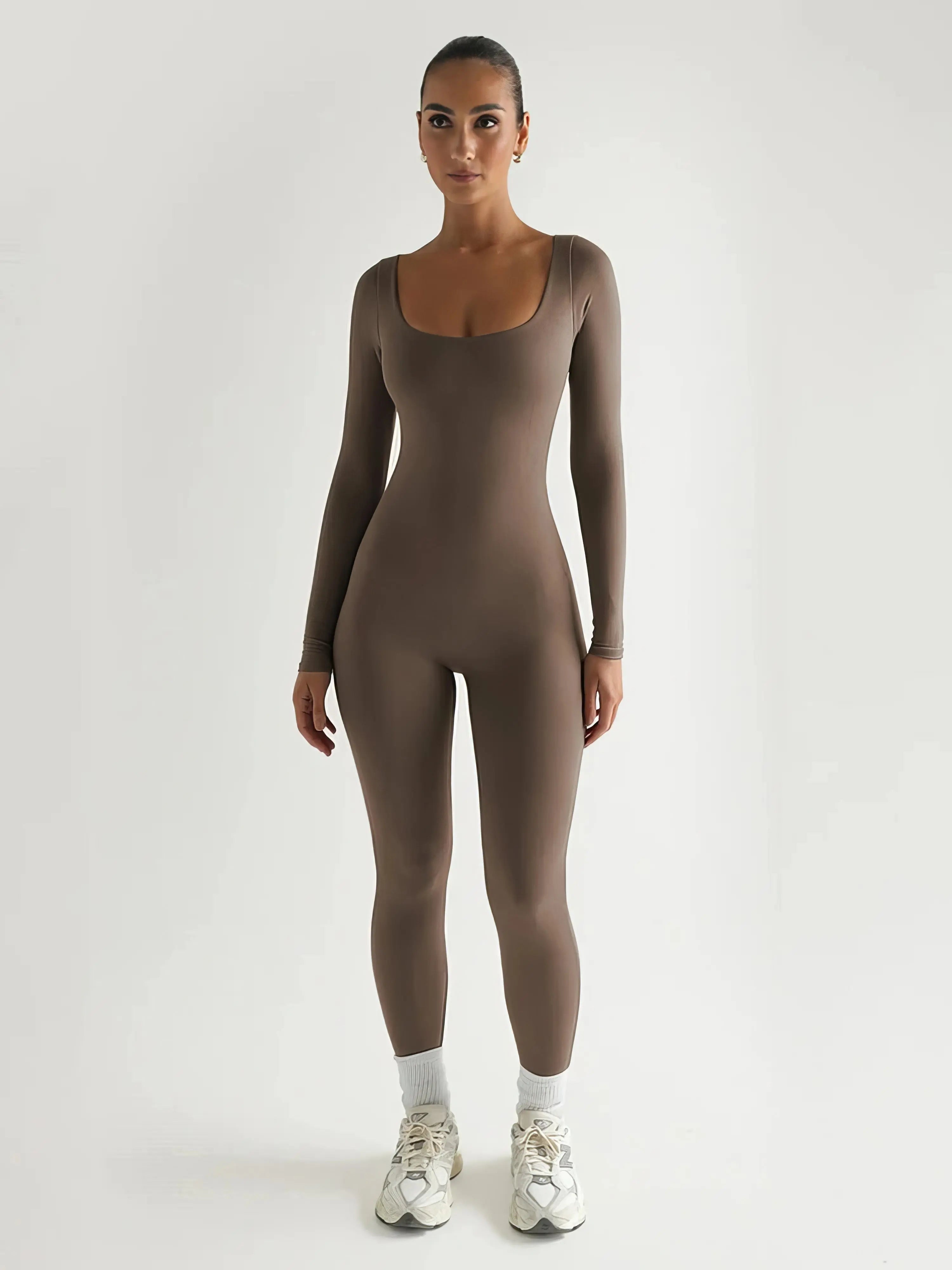 Donna in tuta aderente marrone, maniche lunghe, scarpe da ginnastica bianche, sfondo bianco. Abbigliamento sportivo elegante e moderno.