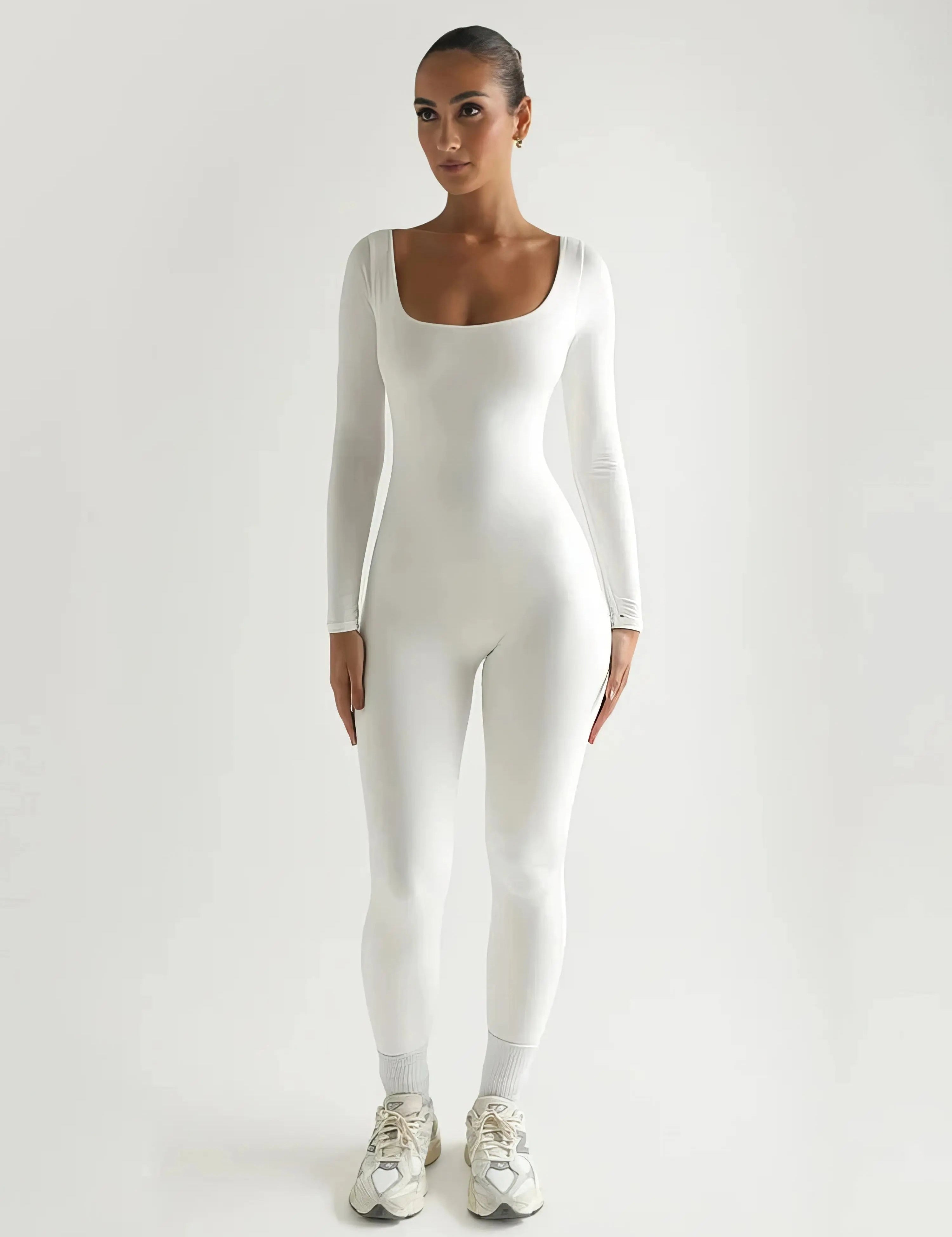 Tuta bianca aderente a maniche lunghe, indossata da una donna in piedi, scarpe da ginnastica bianche, sfondo neutro. Moda donna, abbigliamento sportivo.